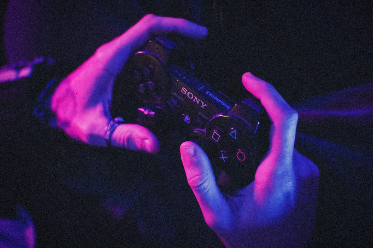 Two hands gripping a black PlayStation 3 controller in a room lit purple and blue.