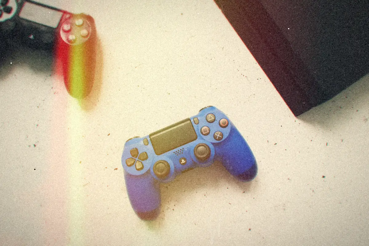 A blue and a black PlayStation 4 controller on a white speckled surface beside the corner of a black PS4.