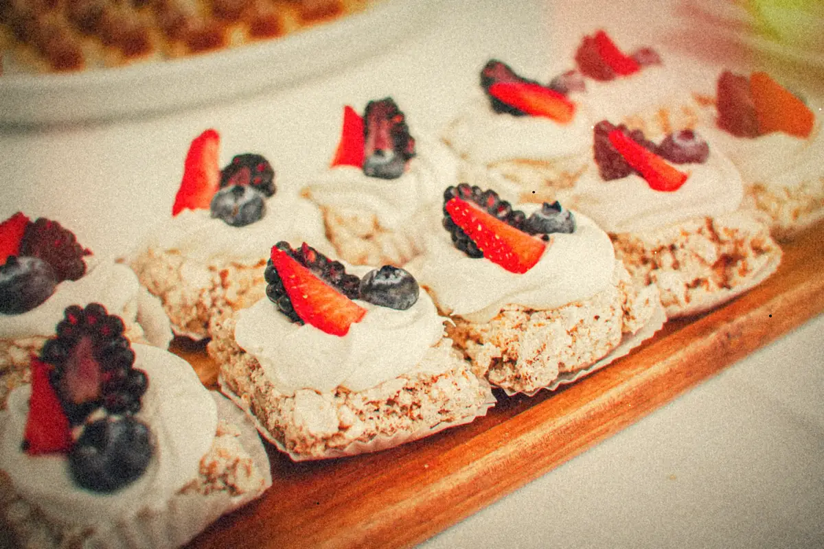 Rows of small cakes topped with whipped cream, sliced strawberries, blackberries and blueberries on a wooden tray.