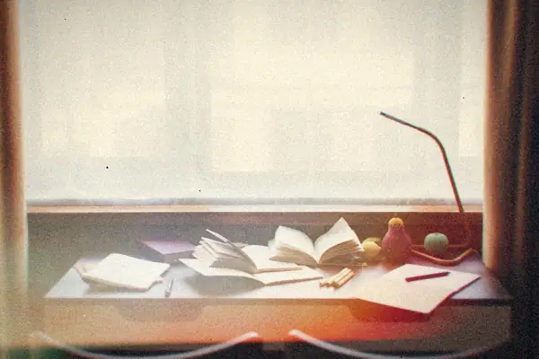 A desk by a curtained window covered with open books, notebooks and a lamp.
