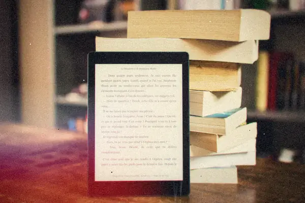 An e-reader showing a page of text propped in front of a stack of paper books.