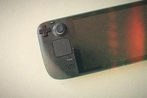 A close-up of a Steam Deck lying on a white surface, showing its d-pad, left stick, trackpad and dark screen.
