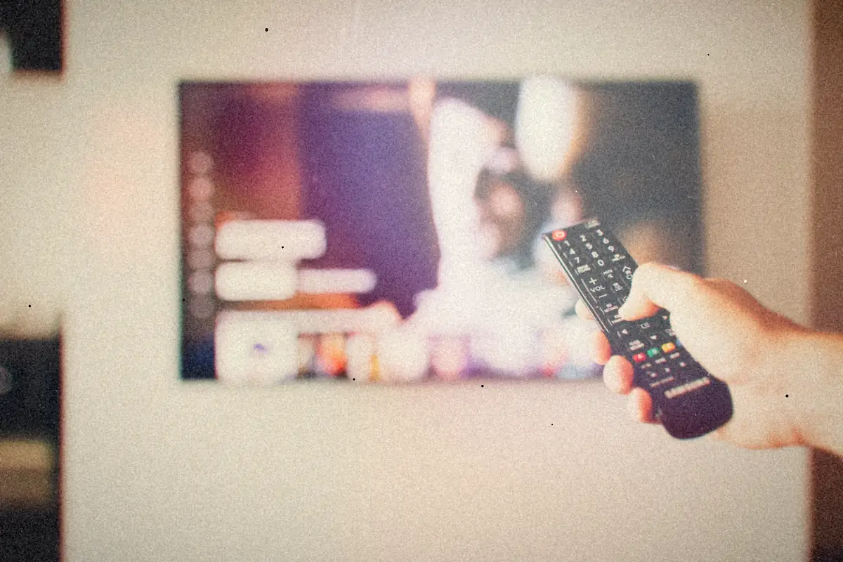 A hand pointing a remote control at a TV showing a streaming menu.
