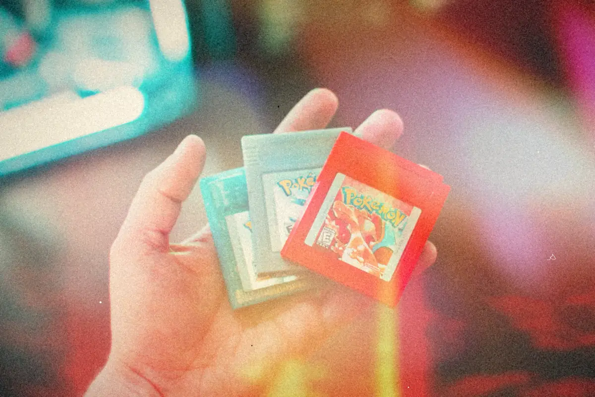 A hand holding three Game Boy cartridges, including a red Pokemon cartridge, in front of a blurry screen.
