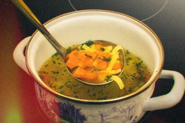 A pot of chicken noodle soup with carrots and parsley, with a ladle lifting out noodles.