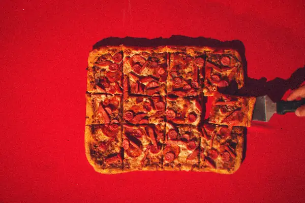 A rectangular pepperoni pizza cut into squares, with one slice being lifted on a spatula against a red background.