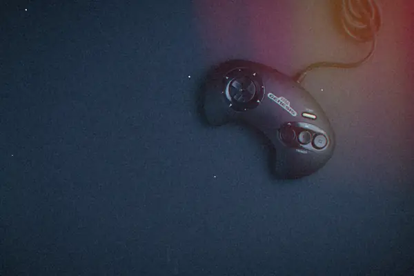 A black three-button Sega Genesis controller lying on a dark navy background, its cord trailing off the top of the frame.
