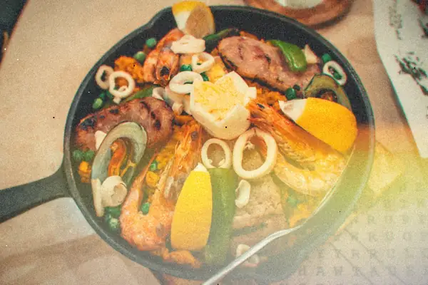 A cast-iron pan of saffron rice with shrimp, grilled sausage, squid rings, peas, peppers and lemon wedges.