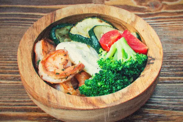 A wooden bowl of seared shrimp with grilled zucchini, broccoli, tomato and a dollop of sauce.