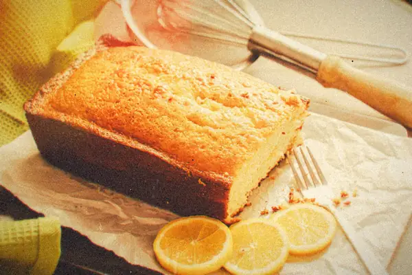 A sliced lemon loaf cake on parchment with lemon slices and a whisk.