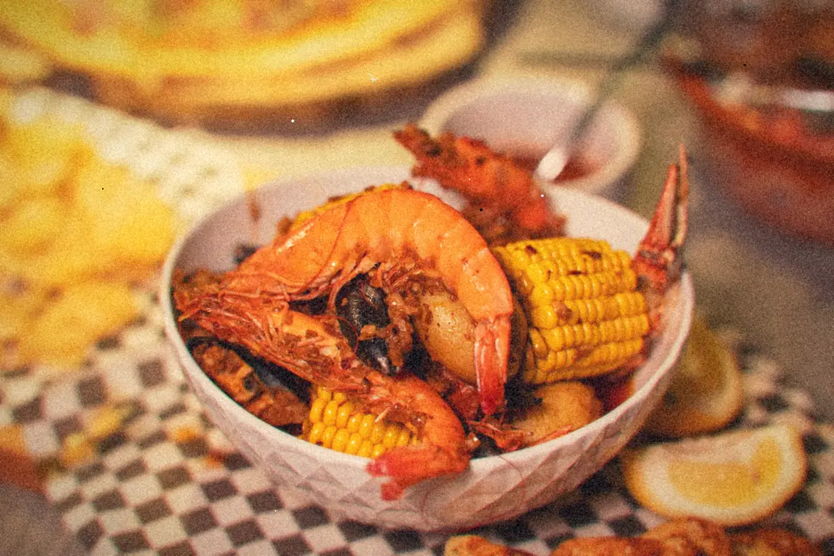 A bowl of seafood boil with whole shrimp, corn on the cob and potatoes beside lemon wedges.