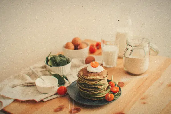 A stack of green spinach pancakes topped with sour cream and cherry tomatoes, with eggs, spinach and milk on a wooden table.