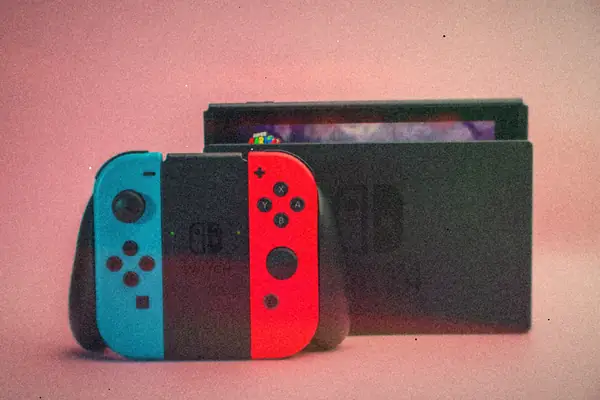 A pair of blue and red Joy-Con controllers in their grip in front of a docked Nintendo Switch on a pink background.