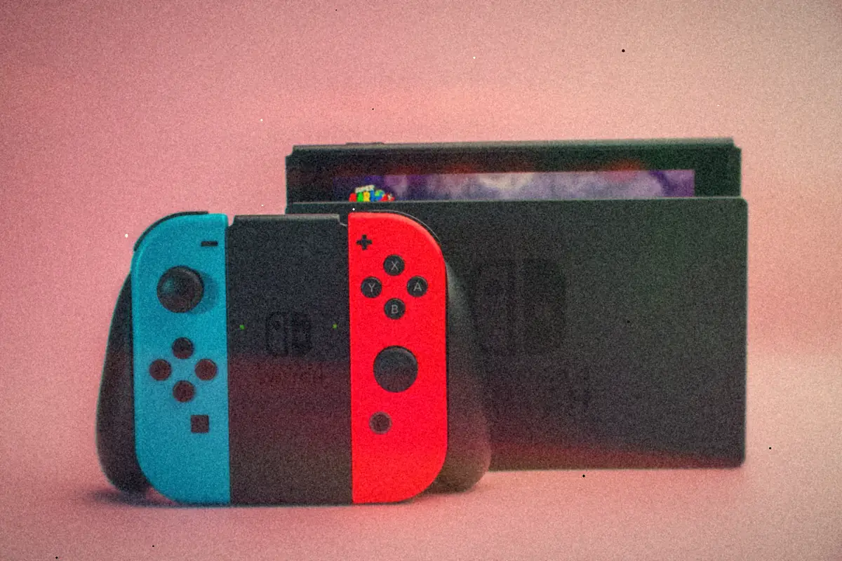 A pair of blue and red Joy-Con controllers in their grip in front of a docked Nintendo Switch on a pink background.