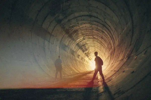 Two people silhouetted in a dark industrial tunnel with light at the far end.