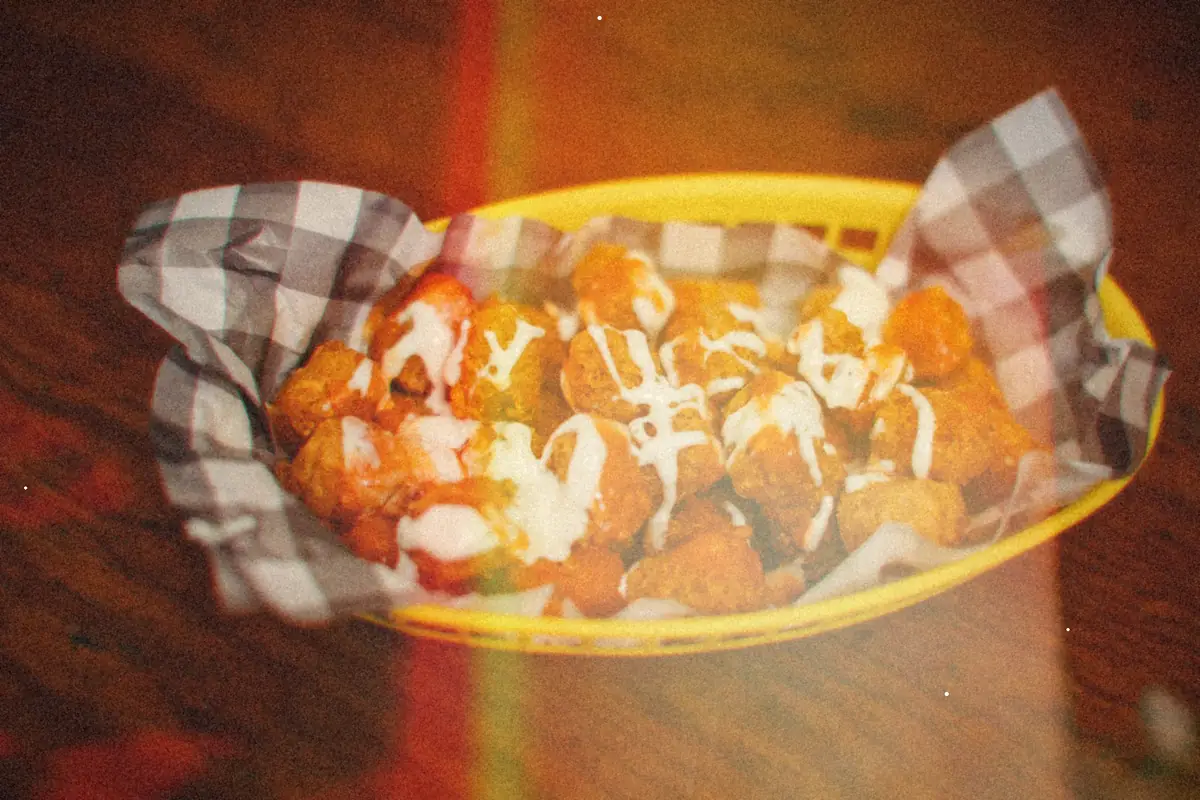 A yellow basket of crispy tater tots topped with cheese and a ranch drizzle on checkered paper.