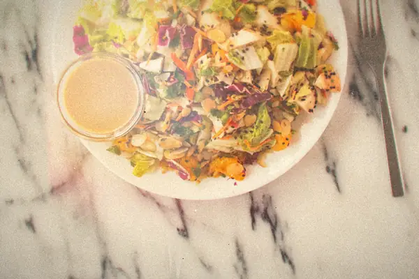 A chopped cabbage salad with carrots, almonds and mandarin oranges beside a cup of peanut dressing.