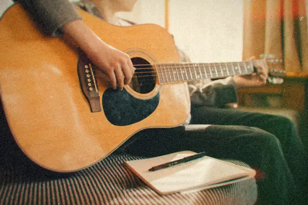 Someone playing an acoustic guitar with a notebook and pen close by.
