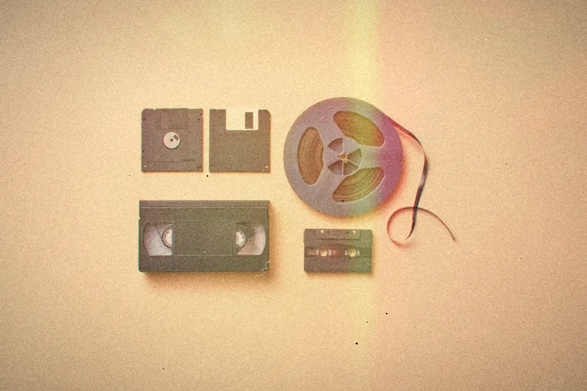 A floppy disk, a VHS tape, a cassette and a reel of tape arranged on a peach background.