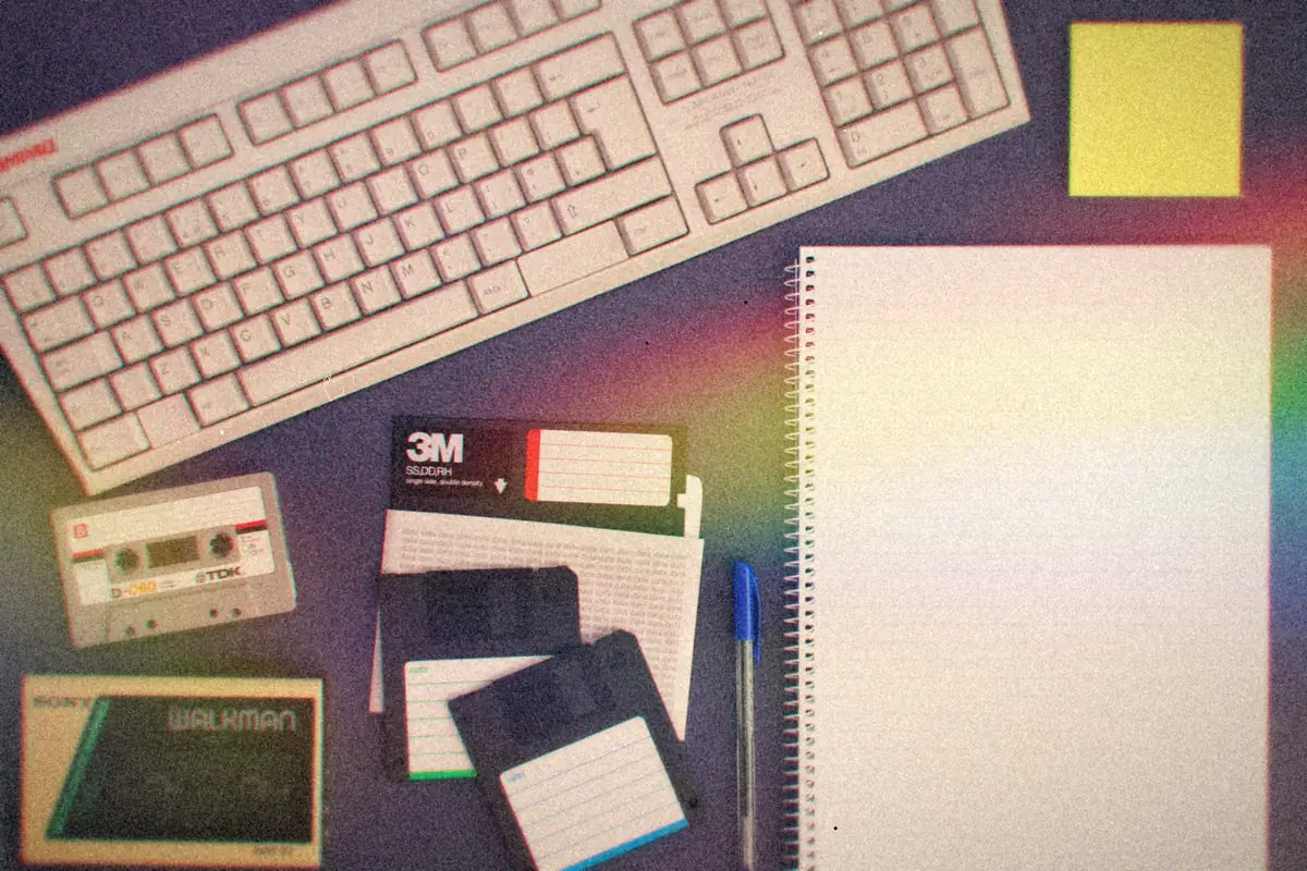 A beige keyboard, floppy disks, cassette tapes, a Walkman and a spiral notepad laid out on a desk.