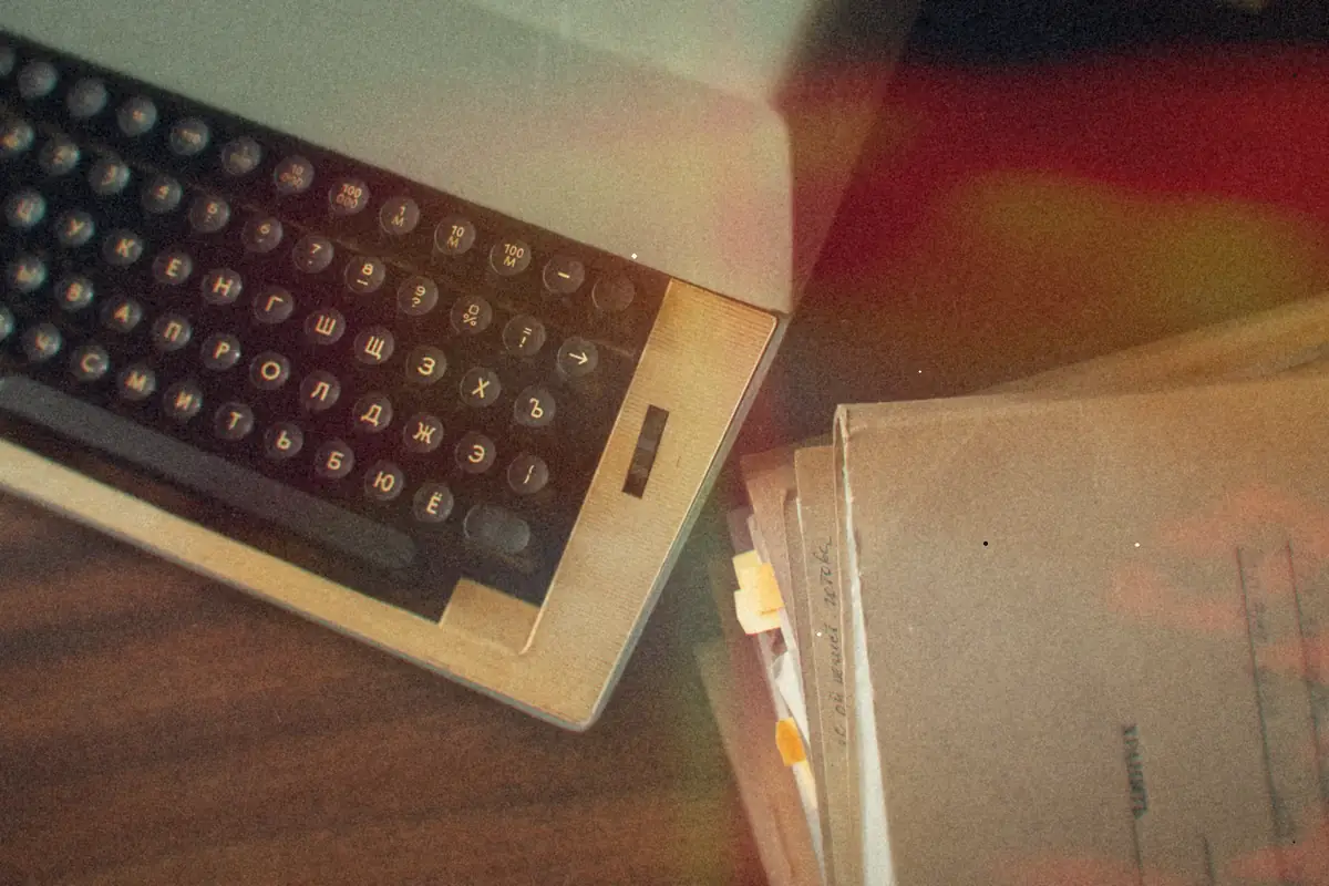 An old typewriter on a wooden desk next to a stack of paper manuscripts and folders.