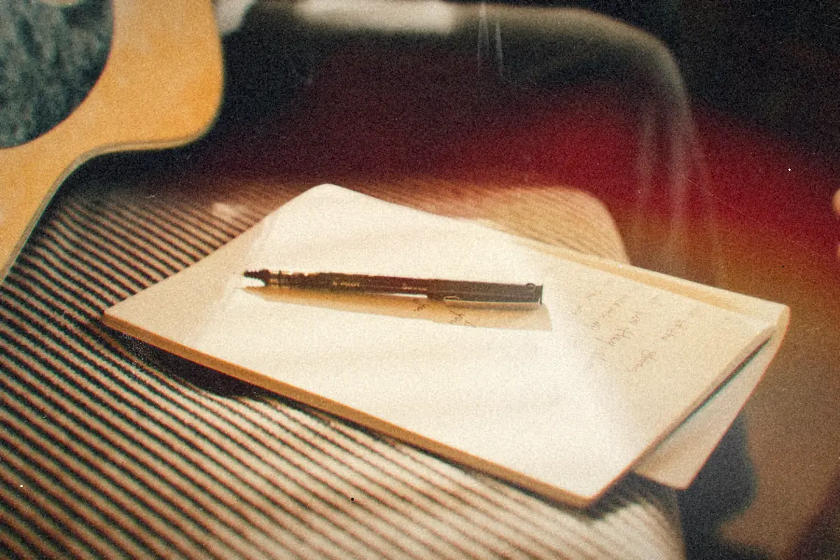 A notebook with handwritten lines and a pen resting beside an acoustic guitar.