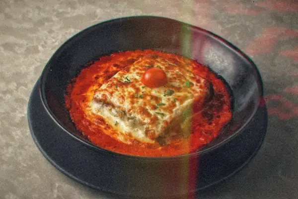 A piece of cheesy baked lasagna sitting in rich tomato sauce in a black bowl, topped with a cherry tomato.