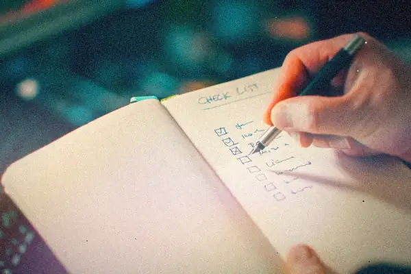 A hand ticking off items on a handwritten checklist in an open notebook, with a laptop behind it.