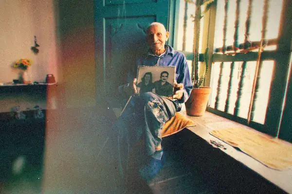 An older man sitting by a window, looking at an old photograph.