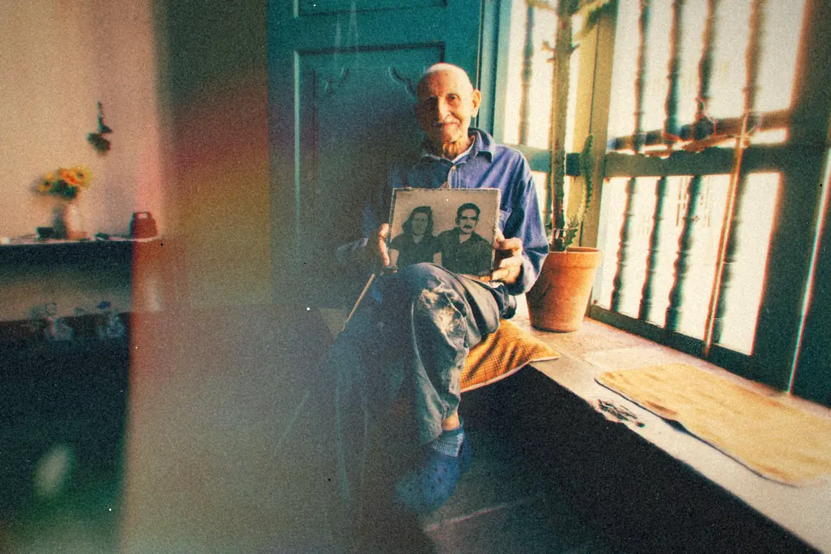 An older man sitting by a window, looking at an old photograph.