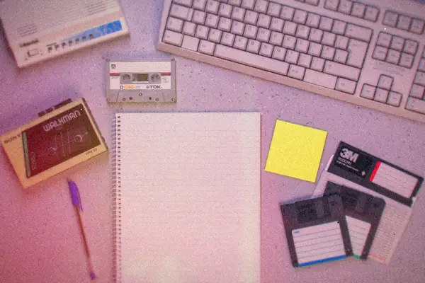 A beige keyboard, a small stack of 3.5-inch floppy disks, a cassette tape and a Walkman laid out on a desk around a blank notepad.