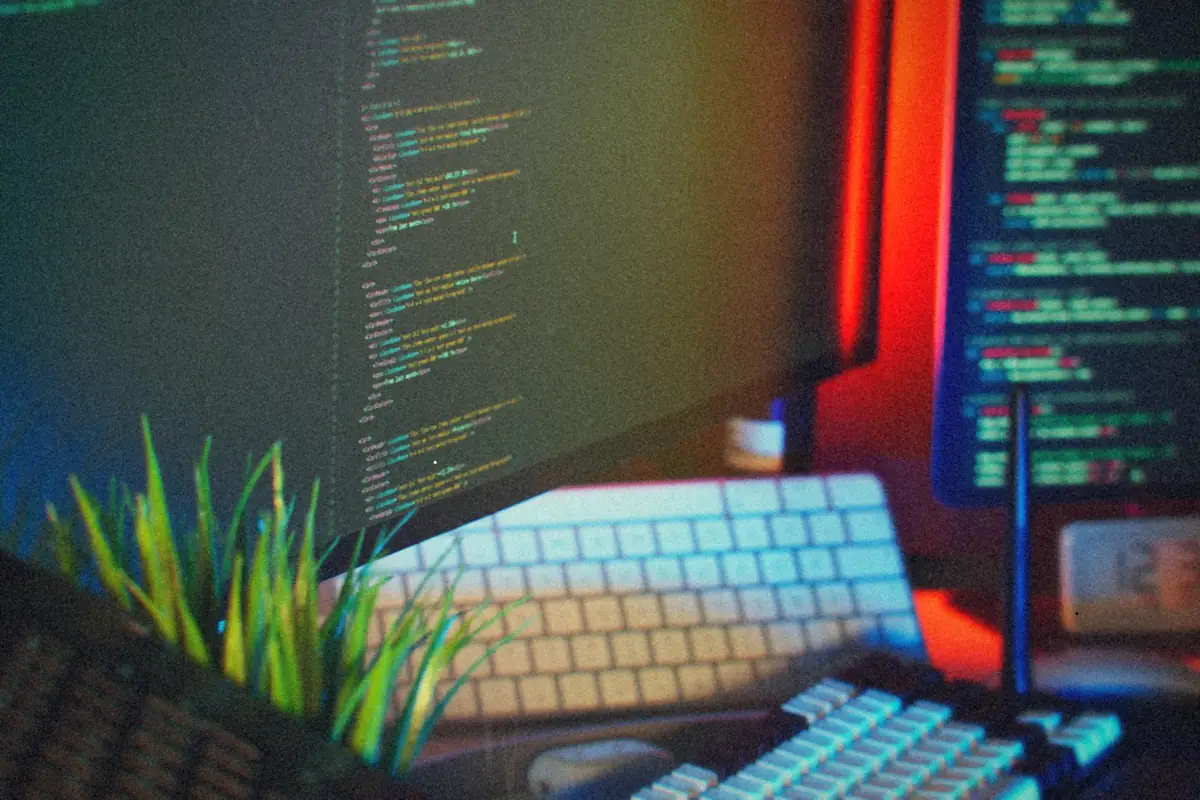 A developer's desk with code on two monitors, a mechanical keyboard, a small clock and a desk plant.