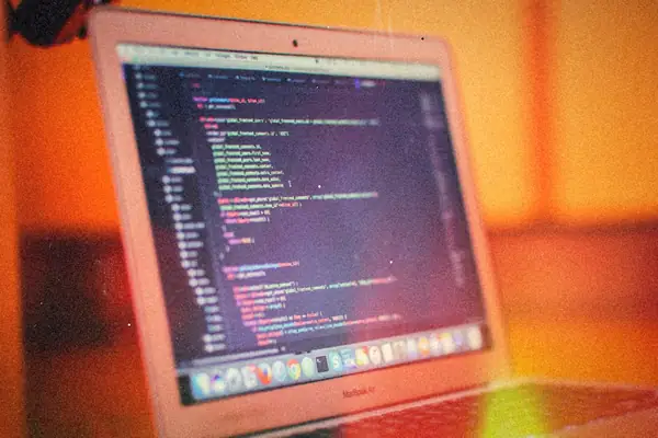 A laptop on a desk showing lines of code in a dark editor, with warm orange light behind it.