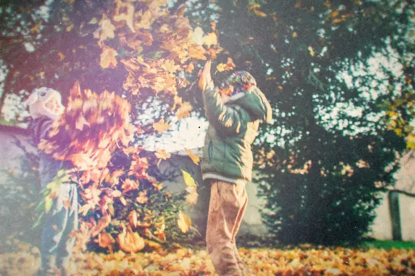 Two kids throw armfuls of fallen autumn leaves in a sunny backyard.