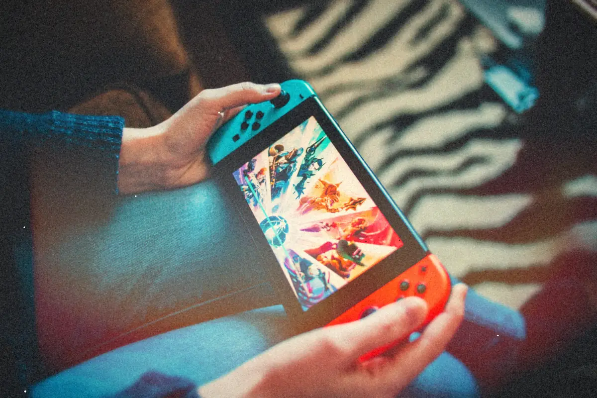 Two hands holding a Nintendo Switch in handheld mode, with blue and red Joy-Cons and a colorful game menu on the screen.
